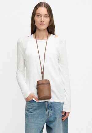 Frau mit langen braunen Haaren, die ein weißes Langarmoberteil, eine blaue Jeans und ein braunes Lederhalsbeutel mit Reißverschlusstaschen trägt.