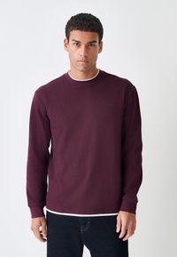 Bordeauxrotes Langarmshirt mit Rundhalsausschnitt und gerippten Bündchen, aus strukturiertem Stoff, über einem weißen T-Shirt getragen, kombiniert mit einer dunklen Jeans.