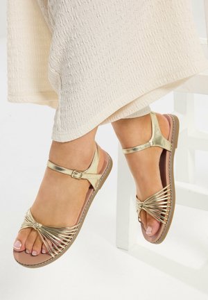 Füße, die goldene flache Sandalen mit Riemchen und Knöchelriemen tragen, ruhen neben einem strukturierten, cremefarbenen Stoff vor einem weißen Hintergrund.