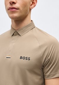 Gros plan d'une personne portant un polo beige avec "BOSS" imprimé sur la poitrine et un col fin perforé.