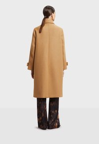 Kaput s odavzanom kroju od kamilje vune, visokog ovratnika i razdvojenih manžeta; upareni s razdivljenim, uzorkovanim tamnim hlačama.