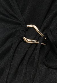 Crni materijal s naboranom teksturom i zlatnim metalnim naglaskom koji podseća na udicu. Dizajn sadrži detalj čvora koji je vezan.