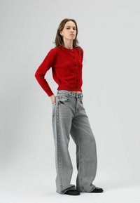 Cardigan rouge en maille avec des boutons noirs, associé à un jean gris ample et des chaussures plates noires. Le mannequin est debout devant un fond neutre.