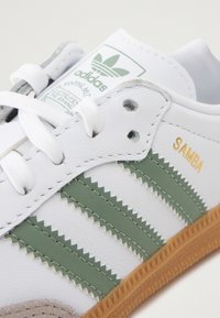 Os ténis Adidas Samba têm um cabedal em pele branca com riscas verdes em zigzag, uma sola de borracha colorida e detalhes em dourado com a inscrição "SAMBA".