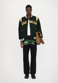 Green varsity jacket with white and yellow accents, featuring "ICE CREAM" lettering. Black pants with neon green graphic. Brown bag in hand.