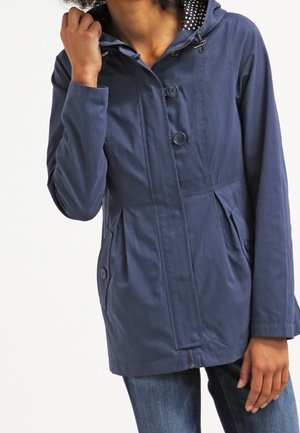 Woman wearing a navy blue buttoned jacket with a hood and polka dot lining, paired with blue jeans, hand near collar.