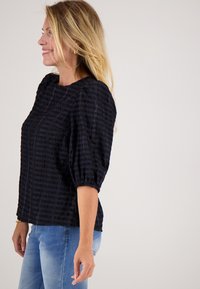 Zwarte blouse met textuur, pofmouwen, ronde halslijn en zijsplitten, gecombineerd met lichtblauwe jeans.