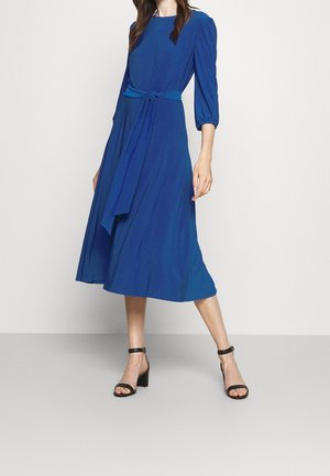 Woman wearing a long-sleeve, knee-length blue dress with a matching belt, paired with black ankle-strap block-heeled sandals, standing against a plain background.