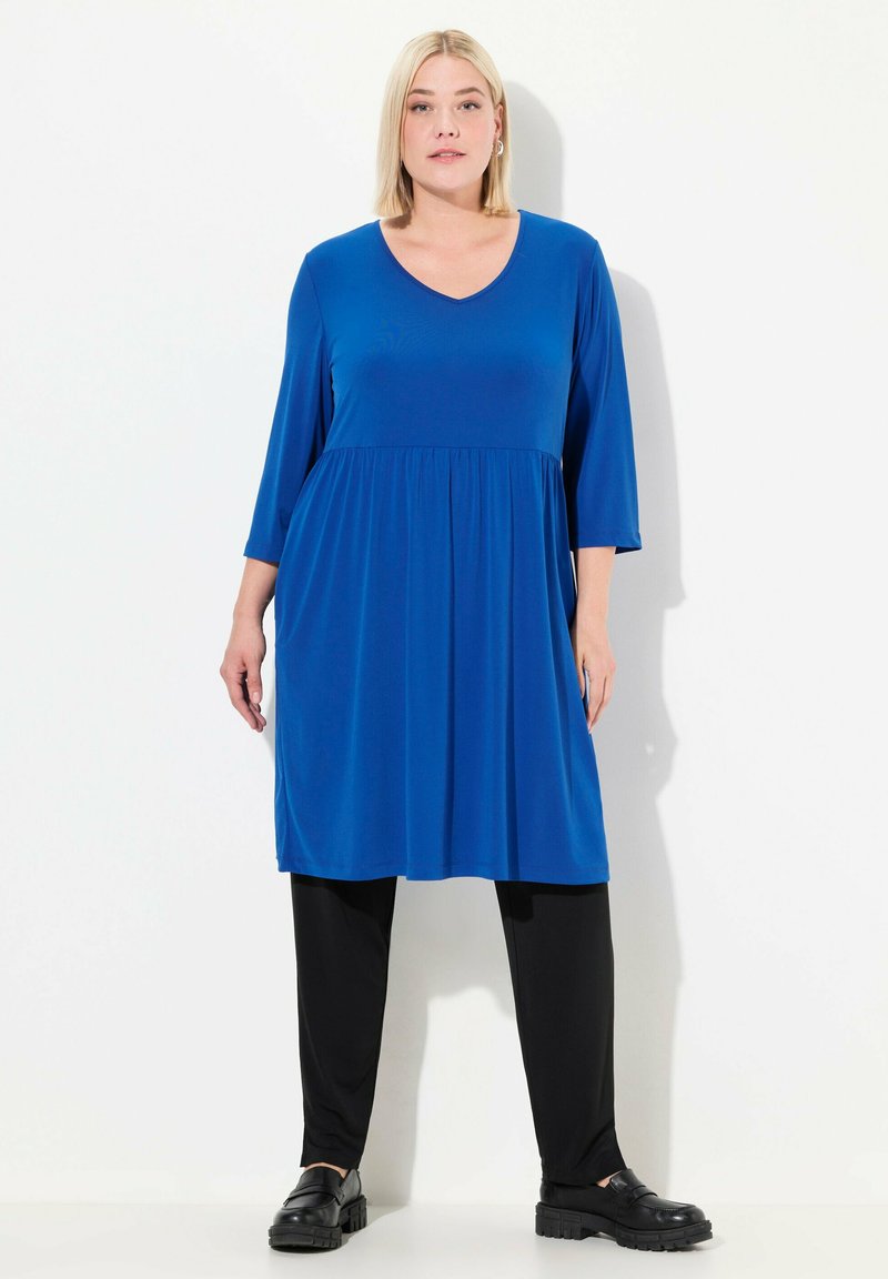 Niebieska sukienka do kolan z dekoltem w kształcie litery V, rękawami do trzech kwart oraz marszczoną talią. Noszona z czarnymi leginsami i czarnymi, masywnymi butami.