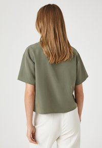 Top de color verde oliva, de manga corta y corte relajado con dobladillo redondeado, fabricado con una tela suave. La vista trasera muestra un diseño simple y líneas limpias.