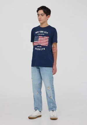 T-shirt in cotone blu navy con grafica dell'American flag invecchiata e testo. Abbinata a jeans strappati blu chiaro e sneaker bianche.