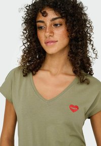 Oliven grøn V-hals t-shirt i blødt stof med korte ærmer og en rød broderet hjerte detalje på venstre bryst.