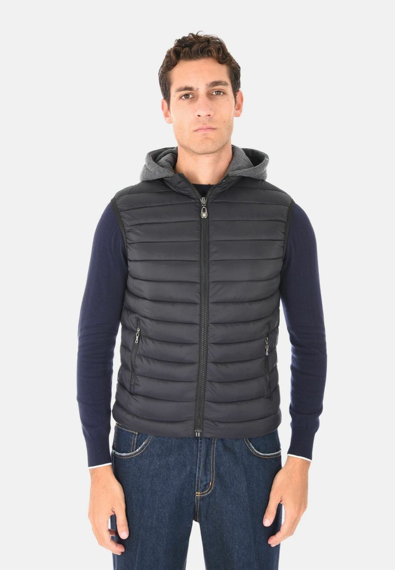 Giovane uomo in piedi, che indossa un maglione blu navy, un gilet senza maniche nero imbottito con cappuccio grigio e jeans blu, rivolto verso l'osservatore su uno sfondo semplice.