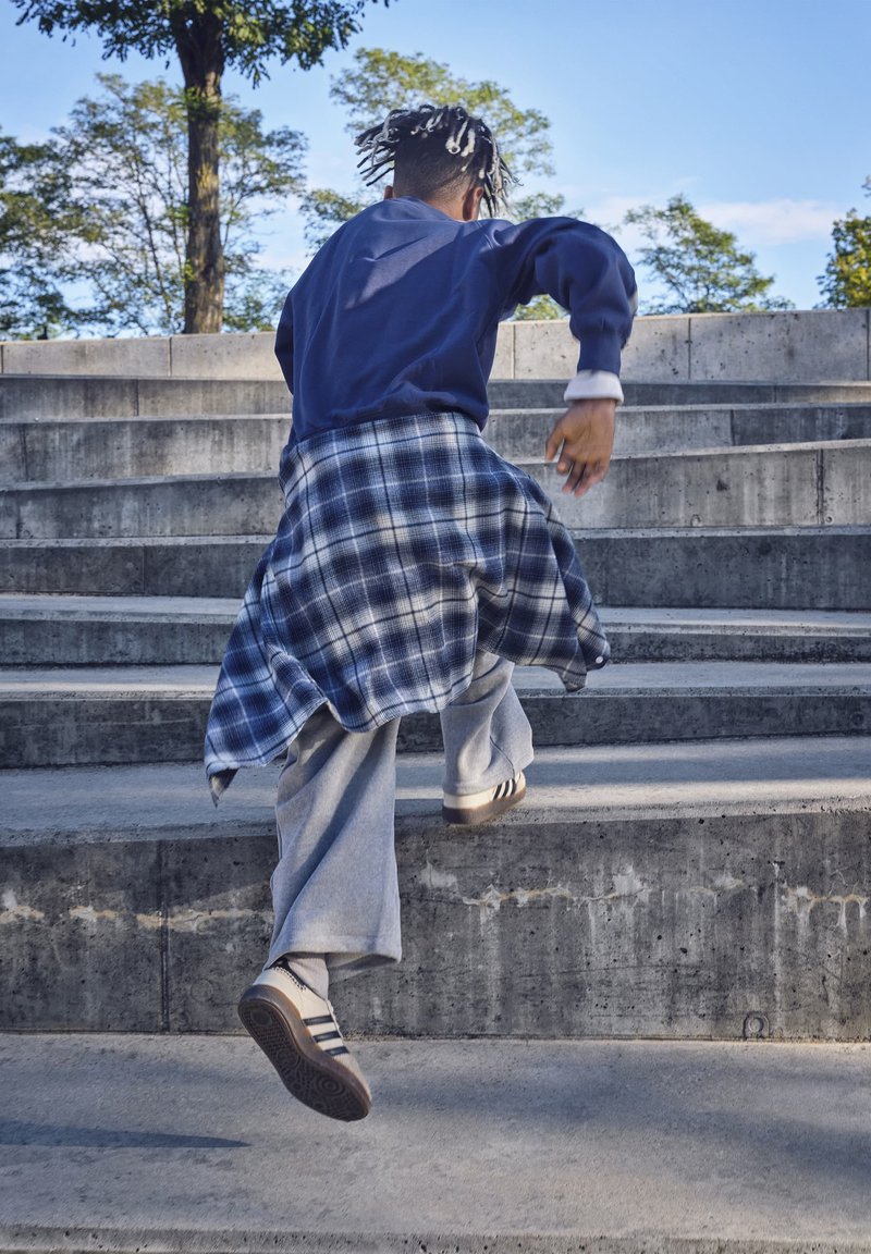 Personne aux cheveux tressés portant une veste bleue et une chemise à carreaux nouée autour de la taille monte des escaliers extérieurs en béton.
