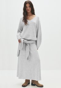 Pulover gri cu guler în V și mâneci lungi, asociat cu o fustă asortată, care are un detaliu de talie legată. Fabricat dintr-un material texturat.
