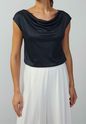 Top azul marino con mangas de capa, confeccionado en tela suave. Combina con pantalones plisados blancos, mostrando una silueta suelta y relajada.