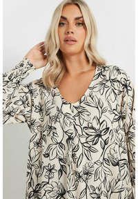 Löst sittande beige blus med V-ringning, prydd med svart blommönster och långa ärmar. Tyget verkar mjukt med en slät yta.