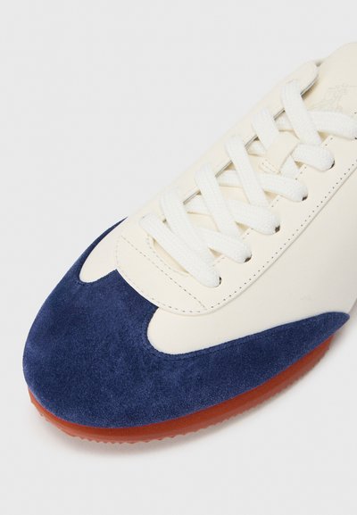 Bőr és velúr sportcipő fehér felsőrésszel, tengerészkék orrkappal, fehér fűzőkkel és narancssárga gumitalppal. Simított és strukturált felület jellemzi.