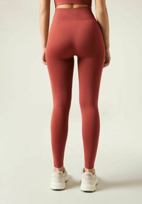 Hochbundige, figurbetonte Leggings aus rostrotem Stoff. Glatte Textur ohne sichtbare Muster oder Akzente. Kombiniert mit hellen Sneakers.