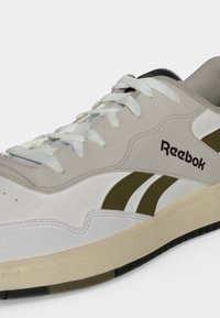 Άσπρο και μπεζ αθλητικό παπούτσι Reebok με ελαιί πράσινες λεπτομέρειες και λευκά κορδόνια, εμφανιζόμενο σε απλό φωτεινό φόντο.