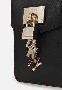 Musta teksturoitu nahkakassi, jossa on kultasävyinen salpalukko ja roikkuvat DKNY-logo-alkuperät kirjaimet, jotka on kiinnitetty suorakaiteen muotoiseen metallilevyyn.