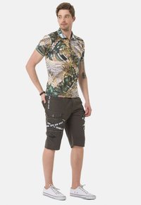 Kurzärmeliges Poloshirt mit einem tropischen Blättermuster in Grün, Braun und Beige, kombiniert mit dunklen knielangen Cargo-Shorts mit weißem Text.