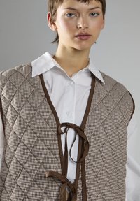 Quiltet vest i brun og beige ternet mønster med brun kant, med V-hals og bånd i taljen, lagdelt over en hvid skjorte med knapper.