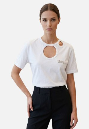 Donna con capelli lisci e scuri che indossa una maglietta bianca con due fori rotondi vicino al colletto e pantaloni neri a vita alta, che posa davanti a uno sfondo semplice.