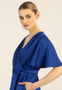 Sinili saten ng balot na damit na may malalapad na flutter sleeves, nakatali sa baywang, at malalim na V-neck. Nagtatampok ito ng makinis na texture at mga side pocket, na nagpapakita ng eleganteng disenyo.