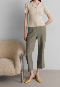 Top corto lavorato a maglia beige con bottoni, pantaloni su misura verde oliva e mule gialli su una modella in piedi accanto a una sedia beige.