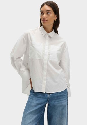 Camicia bianca con bottoni, vestibilità morbida, due tasche sul petto, maniche lunghe e tessuto liscio e leggero. Abbinata a jeans blu.