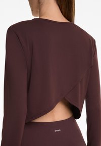 Nainen, jolla on yllään tummanruskea pitkähihainen crop-top, jossa on vino avoin selkä, sekä siihen sopivat korkeavyötäröiset legginsit, joissa on Oysho-logo.