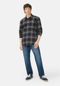 Herren Flanellhemd in schwarz, grau und orange Karomuster; mit Knöpfen und langen Ärmeln. Kombiniert mit blauer Jeans und dunklen Sneakers.