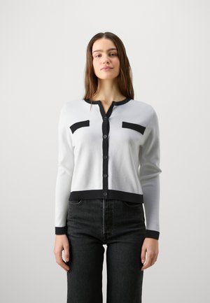 Hvit cardigan med svarte detaljer, med knappelukking foran, lange ermer og to kontrasterende lommer. Ribbestrikket tekstur ved mansjettene og nederkanten.