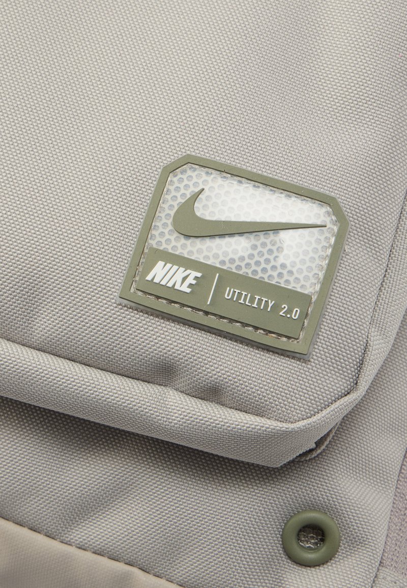 Graues Rucksackmaterial mit strukturierter Oberfläche, versehen mit einem rechteckigen Nike-Label in Grün mit perforiertem Hintergrund und dem Text "Utility 2.0".