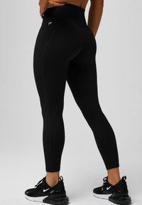 Schwarze, hoch taillierte Leggings aus dehnbarem Material, mit Flachnaht und einem Logo-Akzent. Kombiniert mit schwarzen Sportschuhen.