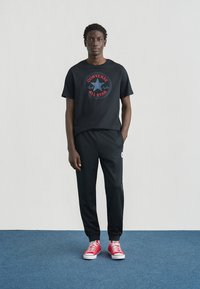 T-shirt en coton noir avec un graphique "Converse All Star" en rouge et blanc, associé à un pantalon de survêtement noir et des baskets en toile rouges.