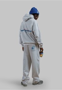 Felpa grigia con testo blu "Leisure Athletics" e pantaloni della tuta abbinati. Presenta un design con tasca ed è abbinata a un berretto blu. Tiene una bevanda.