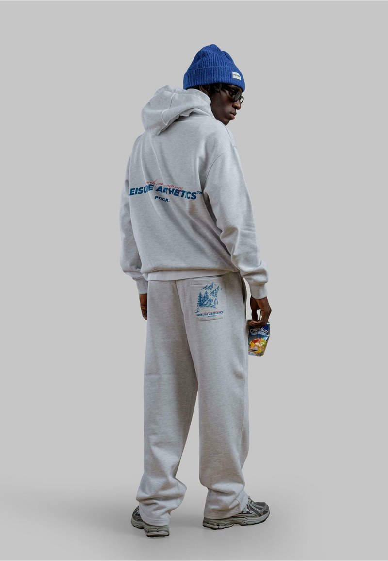 Felpa grigia con testo blu "Leisure Athletics" e pantaloni della tuta abbinati. Presenta un design con tasca ed è abbinata a un berretto blu. Tiene una bevanda.