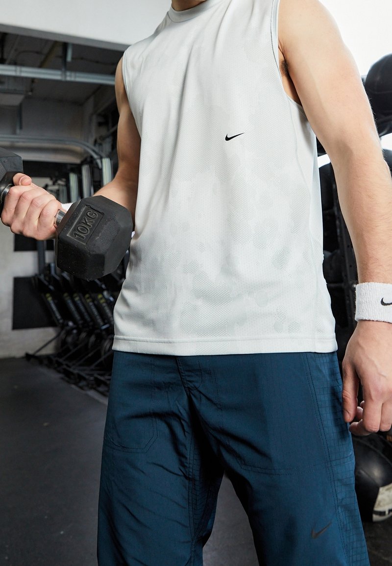 Personne en débardeur blanc et pantalon bleu soulevant un haltère de 10 kg dans une salle de sport.
