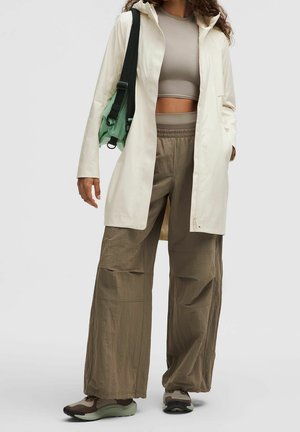 Femme portant un crop top beige, une longue veste crème, un pantalon ample olive, des baskets et un sac vert avec une bandoulière noire sur l'épaule.