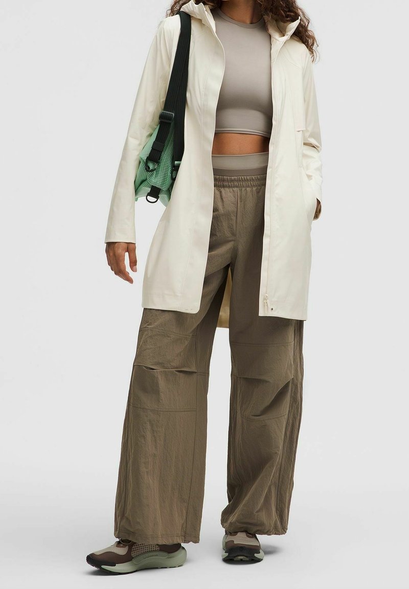 Femme portant un crop top beige, une longue veste crème, un pantalon ample olive, des baskets et un sac vert avec une bandoulière noire sur l'épaule.