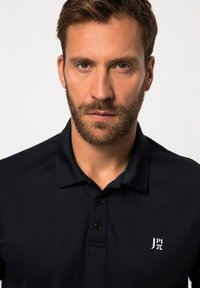 Polo-shirt marine en tissu lisse. Comprend une patte de boutonnage à trois boutons et un petit logo blanc sur la poitrine gauche. Design de col classique.