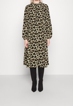 Kobieta w długiej sukience midi z długim rękawem i wysokim kołnierzem w panterkę, w czarnych kozakach za kolano, stojąca na jednolitym tle.