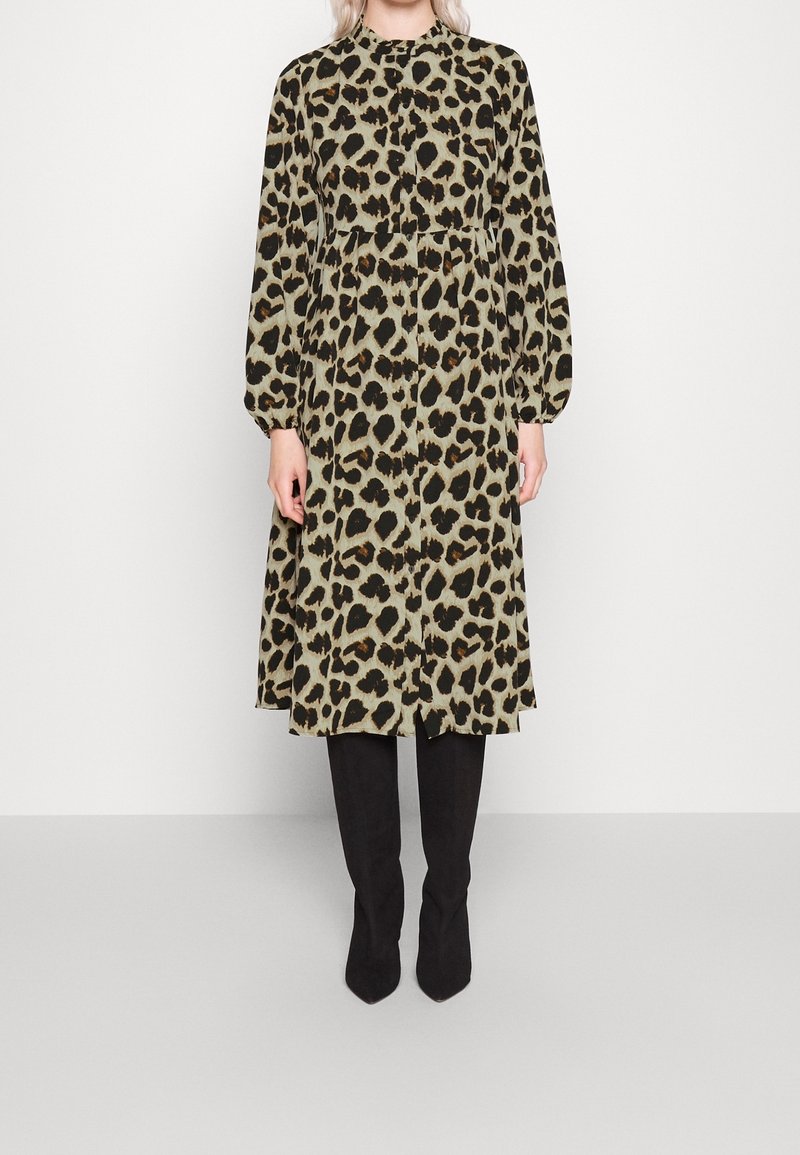 Femme portant une robe midi à manches longues et col montant en imprimé léopard, associée à des bottes noires montantes jusqu'aux genoux, debout devant un fond uni.
