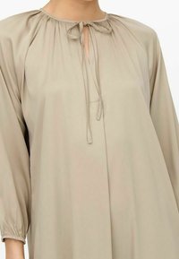 Beige blouse met lange mouwen, gemaakt van lichtgewicht stof, met een sleutelgat-halslijn en een striksluiting, en geplooide details op de schouders.