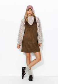 Robe mini en daim marron avec un décolleté en V et des poches latérales, superposée à une chemise à carreaux claire avec des manches volantes ; associée à des chaussures noires brillantes et des chaussettes grises.