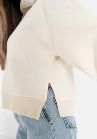 Felpa color crema con fessure laterali e orlo a coste, realizzata in morbido tessuto, abbinata a jeans in denim azzurro chiaro.