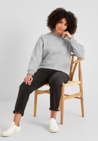 Young woman with curly hair in grey sweatshirt, black rolled-up jeans, and white sneakers sitting relaxed on wooden chair against plain background.
