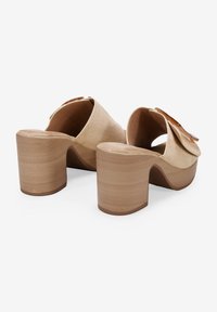 Mules de ante beige con plataforma y parte trasera abierta redondeada. Presentan un tacón de madera grueso y un gran lazo decorativo en la parte superior.